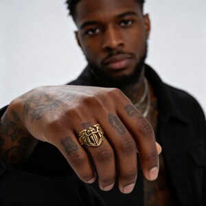 May include: A gold-colored ring with a shield and wing design. The ring features a central emblem with a sword and cross design. The ring is worn on a hand with tattoos.