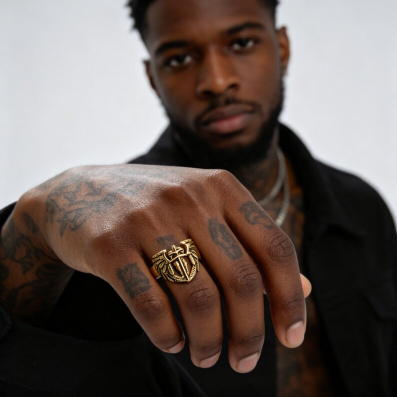 May include: A gold-colored ring with a shield and wing design. The ring features a central emblem with a sword and cross design. The ring is worn on a hand with tattoos.
