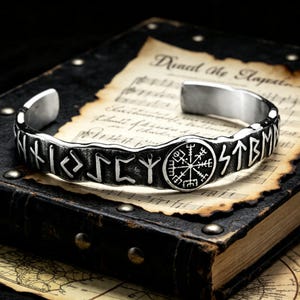 May include: A silver cuff bracelet featuring engraved runes and a central circular emblem. The bracelet is presented on a vintage book with aged paper and musical notation.