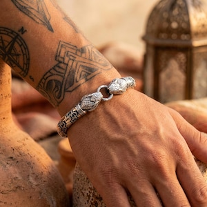 Könnte beinhalten: Ein silberfarbenes Metallarmband mit zwei detaillierten Schlangenköpfen, die an einem zentralen Ring zusammenlaufen. Das Band zeigt ein strukturiertes, sich wiederholendes hexagonales Schuppenmuster mit oxidierten dunklen Akzenten, die einen Kontrast bilden.