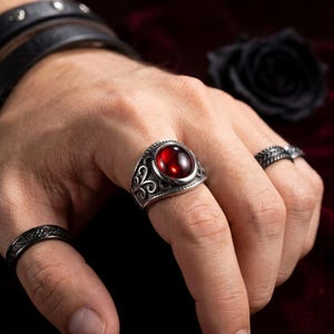 Könnte beinhalten: Ein silberfarbener Ring mit einem großen, glatten, kreisförmigen roten Edelstein, gefasst in eine Ringschiene mit komplexen Schnörkelmustern. Der Ring wird am Mittelfinger getragen, ergänzt durch einen dünnen, strukturierten schwarzen Ring am Daumen und einen schmalen, gemusterten silberfarbenen Ring am Ringfinger.