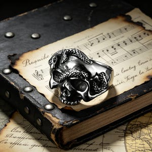 May include: A silver-colored ring featuring a skull design with a snake wrapped around it. The ring is set against a backdrop of an antique book and aged sheet music, creating a gothic or biker aesthetic.