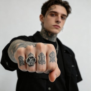 Peut inclure: Une bague argent&eacute;e avec une face octogonale orn&eacute;e d'un motif en relief d&eacute;taill&eacute;. La bague est port&eacute;e sur une main tatou&eacute;e. La personne porte une chemise noire.