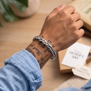 May include: A silver-toned metal cuff bracelet featuring a braided, twisted rope design with an oxidized finish. The bracelet is worn on a wrist, showcasing the intricate, multi-strand woven texture of the metal band against the skin.
