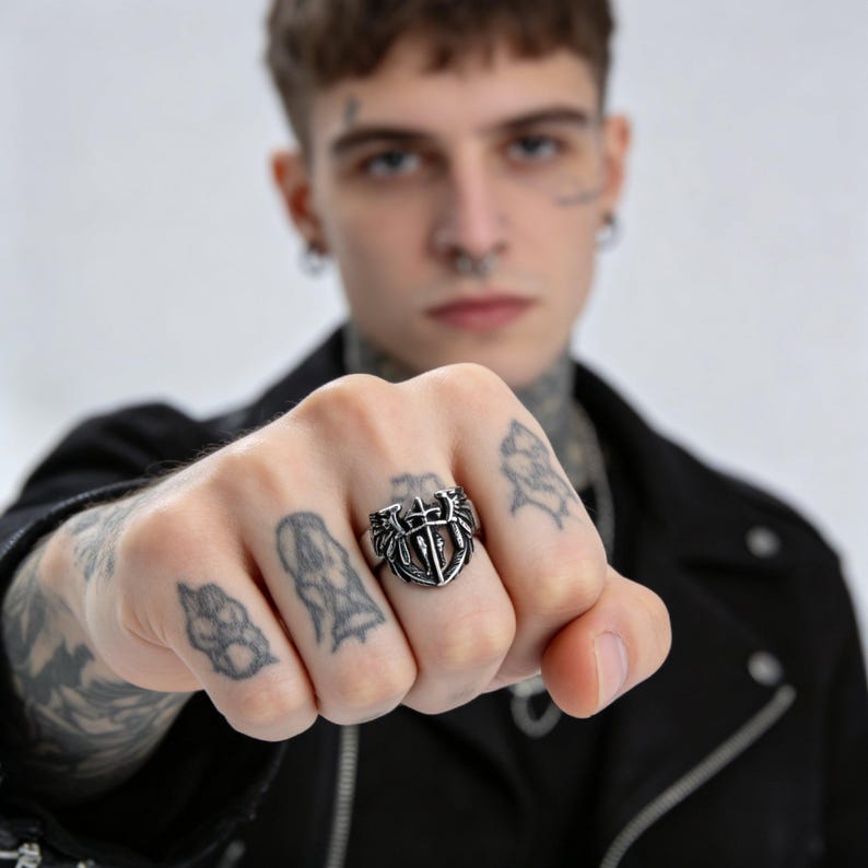 May include: A silver-colored ring with a shield design, featuring a cross and wings. The ring is worn on a hand with visible tattoos. The ring's design is detailed and intricate, with a vintage or gothic aesthetic.