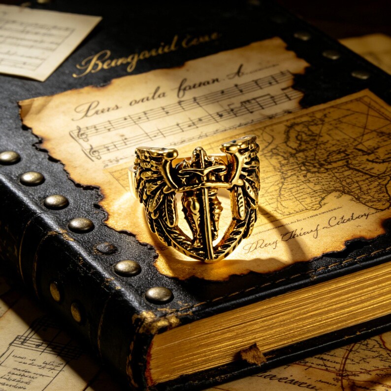 May include: A gold-colored ring with an ornate design featuring a central sword flanked by wings and decorative elements. The ring rests on an open book with aged pages and a map, suggesting a historical or fantasy theme.