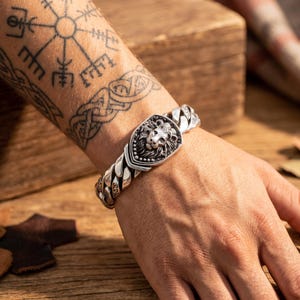 Op de afbeelding: Een zilverkleurige metalen schakelarmband met een centrale schildvormige plaat voorzien van een gedetailleerd leeuwenkopontwerp. De armband bestaat uit dikke, in elkaar grijpende schakels met een gepolijste afwerking en donkere accenten.