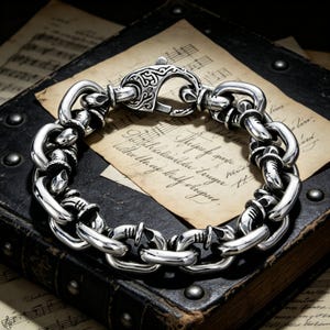 Puede incluir: Una pulsera de cadena plateada con un cierre decorativo. La pulsera presenta eslabones gruesos y redondeados conectados por segmentos texturizados. El cierre tiene un diseño grabado y ornamentado. La pulsera descansa sobre un libro antiguo con partituras.
