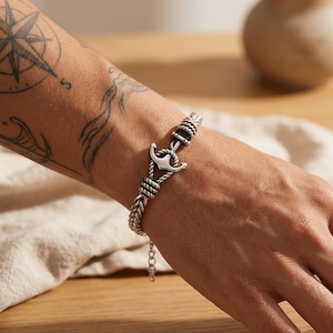 Op de afbeelding: Een zilverkleurige armband met een centrale ankerbedel geïntegreerd in een kettingontwerp met touwtextuur. De armband is voorzien van een kleine bungelende kettingverlenging bij de sluiting en heeft een gedetailleerde metalen afwerking met donkere, geoxideerde accenten.