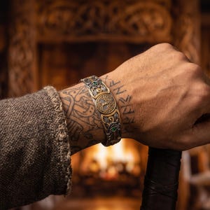 Puede incluir: Un brazalete metálico que presenta un intrincado emblema de árbol en tono dorado en el centro, rodeado de patrones de filigrana en tono plateado y pequeños detalles de piedras preciosas verdes. La pulsera tiene una banda ancha y texturizada.