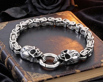 May include: A silver-toned metal bracelet featuring a Byzantine chain link design. The bracelet includes two detailed skull-shaped beads positioned at the front, connected by a circular spring-ring clasp that secures the two ends together.