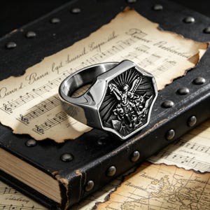 Peut inclure: Bague argent&eacute;e avec une face en forme de bouclier d&eacute;taill&eacute;e repr&eacute;sentant un ange avec des ailes et une &eacute;p&eacute;e. La bague est pos&eacute;e sur un livre ancien en cuir avec des partitions et une carte en arri&egrave;re-plan.