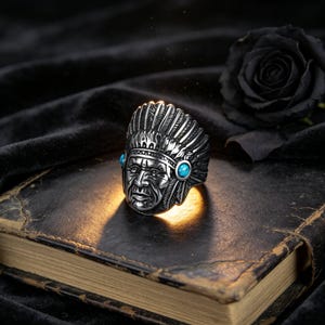 May include: A silver-toned metal ring featuring a detailed relief of a person wearing a feathered headdress. The design includes two small, round, bright blue stones set on either side of the central figure, with oxidized detailing.