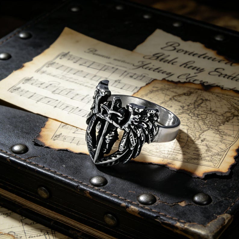 May include: A silver-colored ring with a shield and sword design, set against a backdrop of aged paper and a dark leather surface. The ring features intricate details, including a sword and feather-like wings.