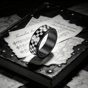 Puede incluir: Un anillo negro y plateado con un patrón de cuadros. El anillo se encuentra sobre un fondo de partituras antiguas y un libro encuadernado en cuero, creando una estética clásica y elegante. El diseño del anillo es una pieza audaz.