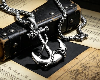 May include: A silver-colored anchor pendant necklace with a rope design wrapped around the anchor. The necklace chain is a box chain style. The background includes sheet music, a map, and a black leather case.
