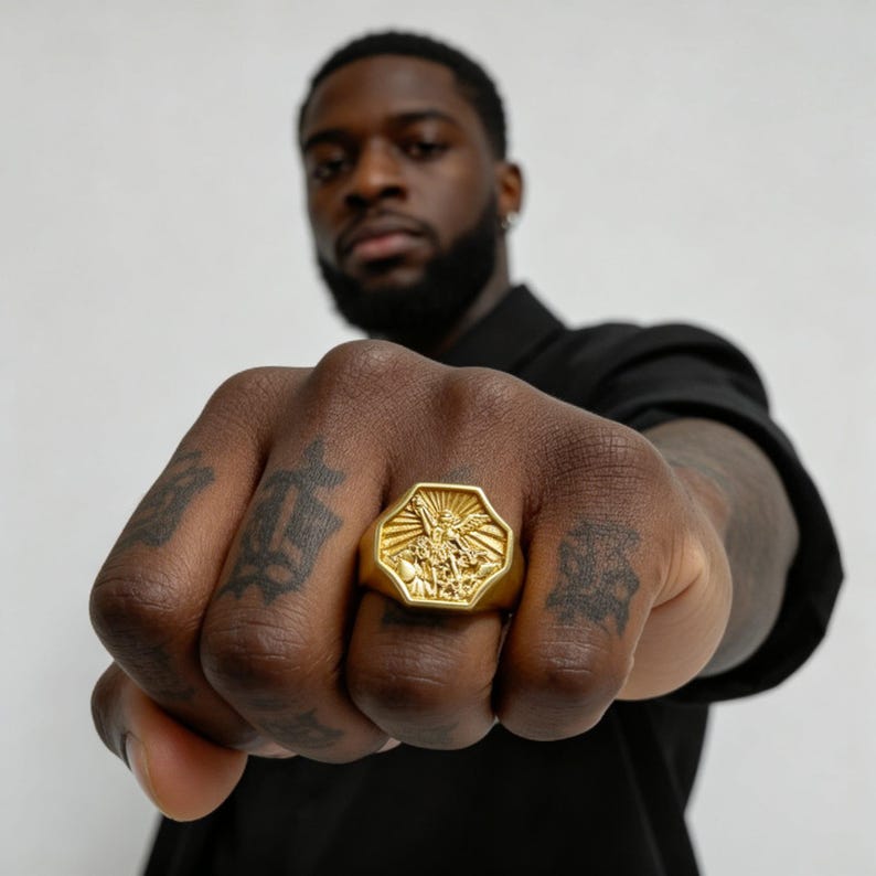 Peut inclure: Une bague chevali&egrave;re dor&eacute;e avec une face octogonale. La bague pr&eacute;sente un motif en relief d&eacute;taill&eacute; d'une figure avec une &eacute;p&eacute;e et des rayons de lumi&egrave;re. La bague est port&eacute;e sur une main tatou&eacute;e.