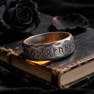 Op de afbeelding: Een zilverkleurige metalen ring met een gehamerd, getextureerd oppervlak en gegraveerde runentekens rondom de buitenkant. De ring heeft een donkere, verweerde afwerking en straalt een warm licht uit vanuit de binnenzijde.