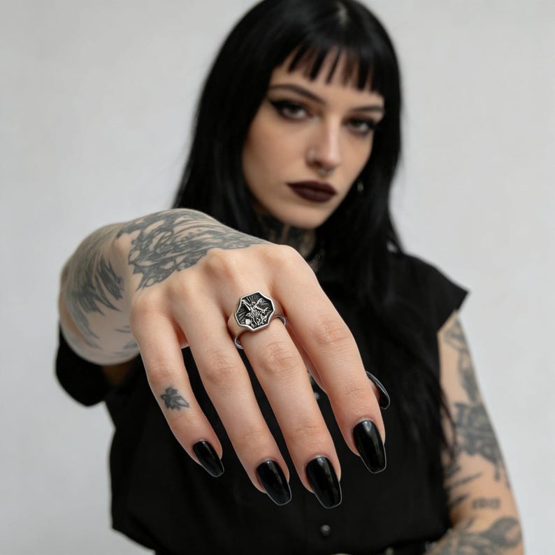 Peut inclure: Bague argent&eacute;e avec une face octogonale orn&eacute;e d'un motif gothique d&eacute;taill&eacute;. La bague est port&eacute;e sur une main avec du vernis &agrave; ongles noir. Le motif de la bague semble &ecirc;tre une image stylis&eacute;e d'une ch&egrave;vre.
