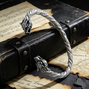 May include: Silver-toned cuff bracelet featuring wolf head terminals. The bracelet has a twisted rope design and is displayed on a vintage leather case with aged paper and musical notes in the background. The bracelet is a bold accessory.