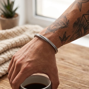 Può includere: Un bracciale a fascia in argento caratterizzato da un dettagliato motivo a spina di pesce intrecciato su tutta la superficie. Il metallo presenta una finitura lucida con aree incassate scure che mettono in risalto la trama complessa.