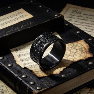 May include: A black ring with engraved runic symbols. The ring is displayed on a vintage sheet of music and a leather-bound book. The ring's design features intricate details and a polished finish, creating a bold and stylish accessory.