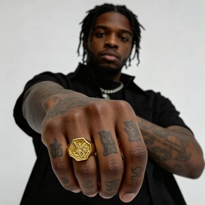 Peut inclure: Bague octogonale dor&eacute;e avec un motif en relief d&eacute;taill&eacute;. La bague est port&eacute;e sur une main avec des tatouages visibles. Le motif de la bague comprend une figure centrale avec des ailes d&eacute;ploy&eacute;es, entour&eacute;e de rayons.