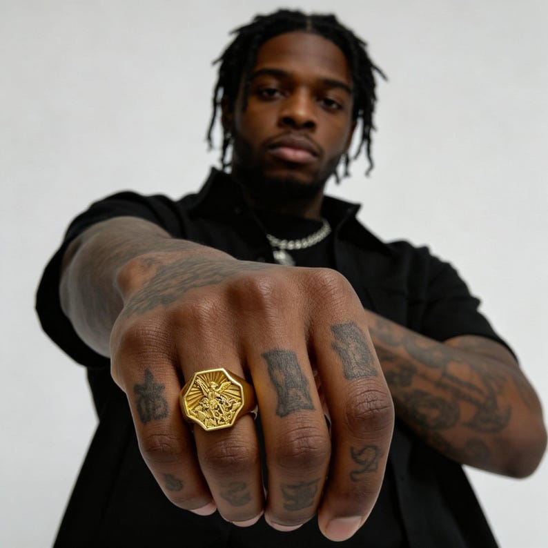 Peut inclure: Bague octogonale dor&eacute;e avec un motif en relief d&eacute;taill&eacute;. La bague est port&eacute;e sur une main avec des tatouages visibles. Le motif de la bague comprend une figure centrale avec des ailes d&eacute;ploy&eacute;es, entour&eacute;e de rayons.