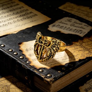 May include: A gold-colored ring with a detailed shield design featuring a sword and crown. The ring is set against a backdrop of aged paper and a leather-bound book, creating a vintage aesthetic. The ring's intricate details are highlighted by the warm lighting.