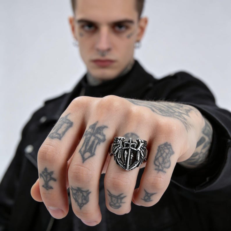May include: A silver-colored ring with a shield design featuring swords and wings. The ring is worn on a hand with multiple tattoos. The ring's design is detailed and intricate, with a gothic or medieval aesthetic.