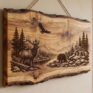 Puede incluir: Una placa de madera rectangular con borde natural que presenta una escena detallada de pirograbado de un paisaje forestal. La obra muestra un alce, un oso, dos lobos y un águila volando entre pinos, un río y montañas, suspendida por una cuerda.