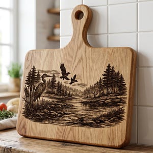 May include: Wooden cutting board with a handle, featuring a detailed engraved nature scene. The design includes a heron, flying birds, trees, and a river. The board is made of light-colored wood.