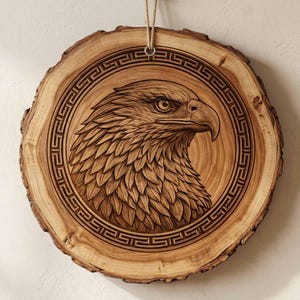 Puede incluir: Adorno de pared de madera circular con un tallado de la cabeza de un águila. El águila está dentro de un círculo con un borde geométrico. La madera es de color marrón claro con un borde texturizado.