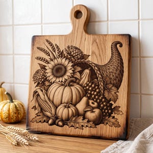 May include: A square wooden cutting board with a handle featuring a detailed laser-engraved cornucopia design. The artwork displays a harvest arrangement of pumpkins, sunflowers, grapes, pears, wheat stalks, and acorns on the light wood surface.
