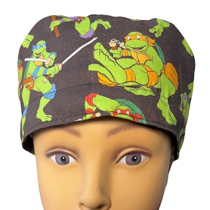 Puede incluir: Un gorro quirúrgico gris oscuro con un patrón repetido de personajes de las Tortugas Ninja verdes, naranjas, azules y rojos. El gorro está diseñado para cubrir la cabeza y el cabello, comúnmente utilizado en entornos médicos.