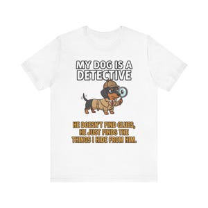 Op de afbeelding: Wit T-shirt met een cartoon teckel detective met een trenchcoat en hoed, die een vergrootglas vasthoudt. Tekst luidt "MY DOG IS A DETECTIVE" en "HE DOESN'T FIND CLUES, HE JUST FINDS THE THINGS I HIDE FROM HIM."