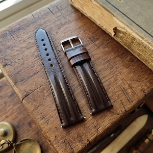 May include: A dark brown leather watch strap with a silver buckle. The strap has visible stitching along the edges and several holes for adjustment. The leather has a glossy finish, and the strap is displayed on a wooden surface.