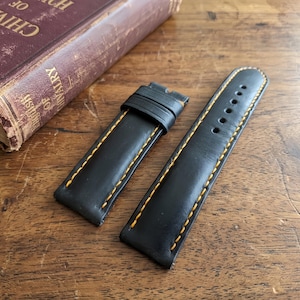 Op de afbeelding: Twee zwarte leren horlogebandjes met gele stiksels. Eén bandje heeft een gesp en de andere heeft meerdere gaten. De bandjes liggen op een houten oppervlak naast een boek met gouden letters.