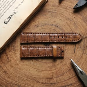 May include: A two-piece brown leather watch strap featuring a textured alligator-style embossed pattern. The strap includes visible stitching along the edges, a single leather keeper loop, and pre-punched holes for adjustment on the longer section.