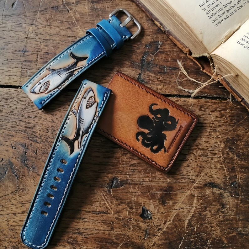 May include: Blue leather watch straps with a shark design and a brown leather cardholder with an octopus design. The watch straps have a silver buckle and white stitching. The shark design is detailed with white and gray accents.
