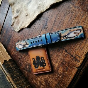 May include: Blue leather watch band with shark illustrations. The band is detailed with a blue buckle and stitching. A brown leather cardholder with an octopus design is also visible.