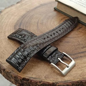 May include: A dark brown watch strap with a glossy, embossed alligator skin texture. The strap features a series of adjustment holes and a polished silver-toned metal buckle. The design includes a matching leather keeper loop for securing the end.