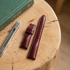 May include: A two-piece dark burgundy leather watch strap with matching stitching. The strap consists of a shorter piece with two leather loops and a longer piece with a tapered end. The leather has a smooth, polished finish and a firm structure.
