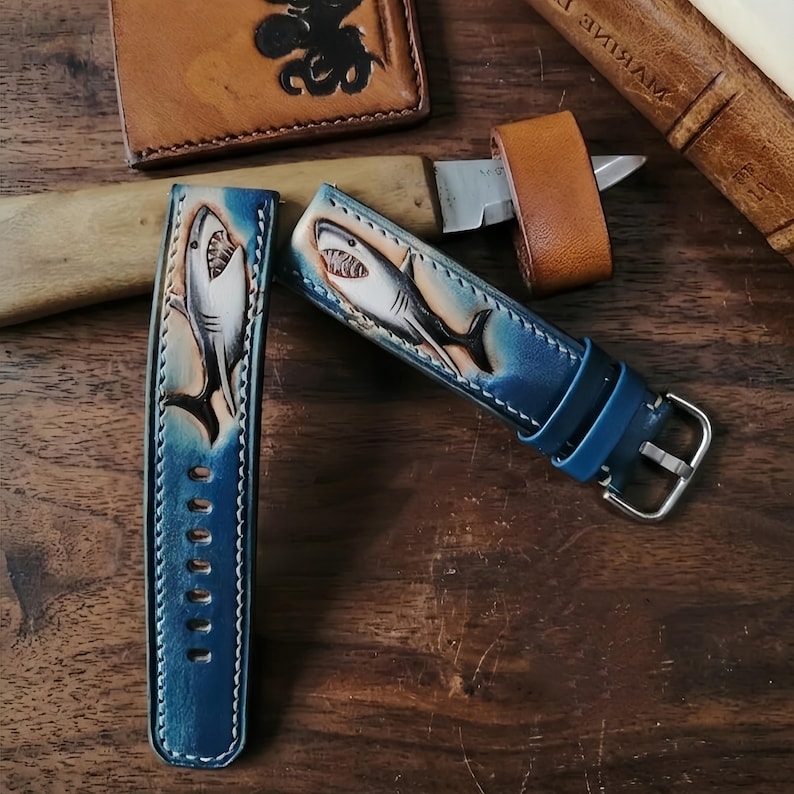 May include: Two blue leather watch straps with a shark design. The straps are hand-tooled and painted with a gradient of blue and white. One strap is fastened with a silver buckle. The straps are displayed on a wooden surface.