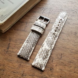 May include: A pair of watch straps with a snakeskin pattern in shades of gray and white, featuring a silver buckle. The straps are displayed on a wooden surface next to a vintage book.