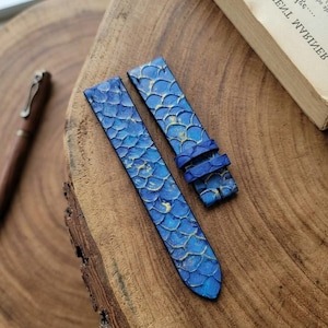 Könnte beinhalten: Ein zweiteiliges Uhrenarmband mit einem blau-goldenen Schuppenmuster. Das Armband hat eine strukturierte Oberfläche mit überlappenden, abgerundeten Formen in verschiedenen Blautönen und metallischen Goldakzenten zur Befestigung an einer Uhr.