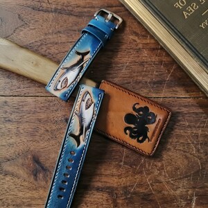 May include: Blue leather watch straps with a shark design and a brown leather cardholder with an octopus design. The watch straps have a silver buckle and are on a wooden surface.