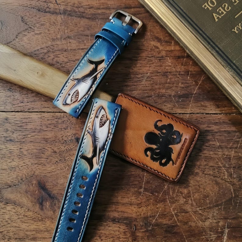 May include: Blue leather watch straps with a shark design and a brown leather cardholder with an octopus design. The watch straps have a silver buckle and are on a wooden surface.