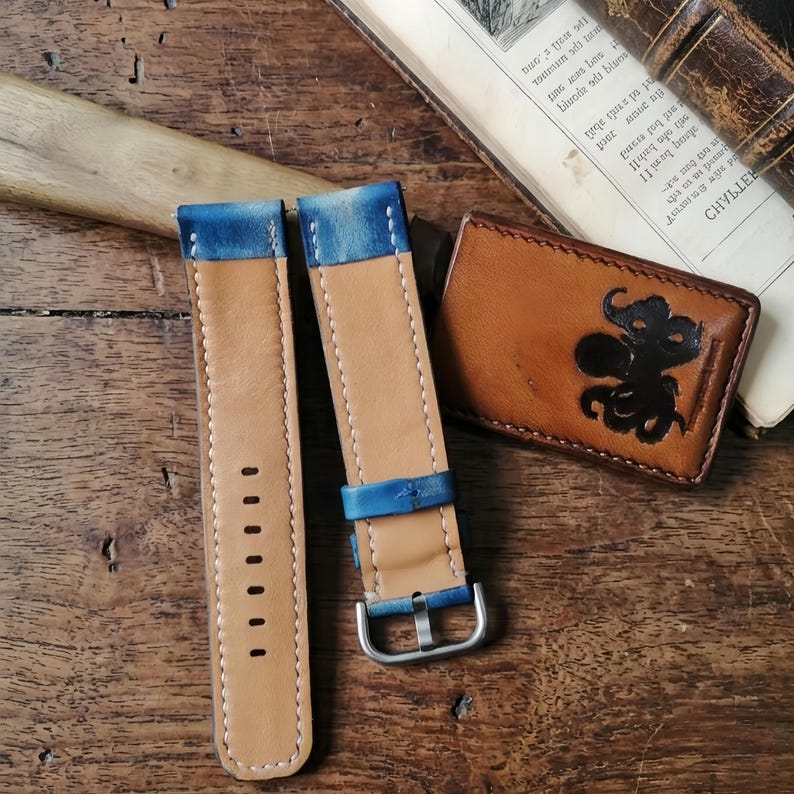 May include: Two leather watch straps with blue accents and a silver buckle. The straps are light brown with blue edges. A brown leather wallet with a black octopus design is also visible.