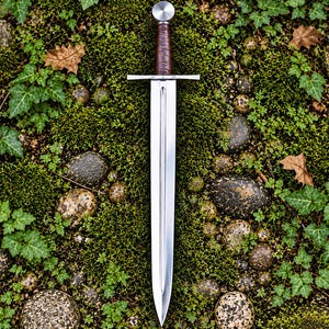 May include: A double-edged steel sword featuring a central fuller, a straight crossguard, and a brown leather-wrapped grip topped with a circular metal pommel. The blade has a polished finish and tapers to a sharp point at the bottom.